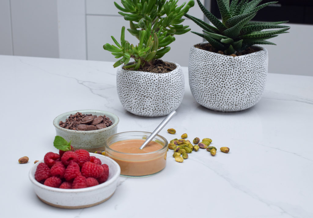 A marble kitchen counter top, with a bowl of raspberries, a bowl of chocolate chips, a pot of peanut butter and two house plants.